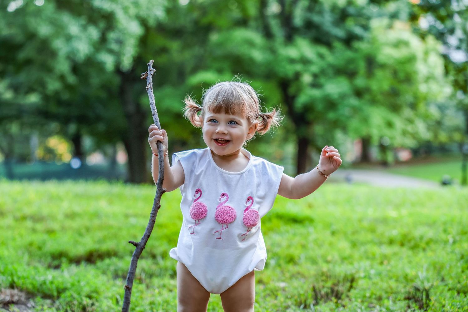 Happy toddler girl in flamingo onesie holding a stick while playing outdoors in green park.