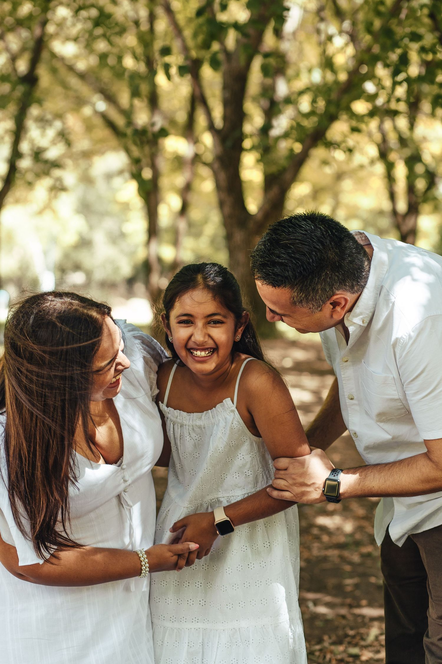 A joyful family shares a candid moment outdoors in matching white outfits on a sunny day among trees.