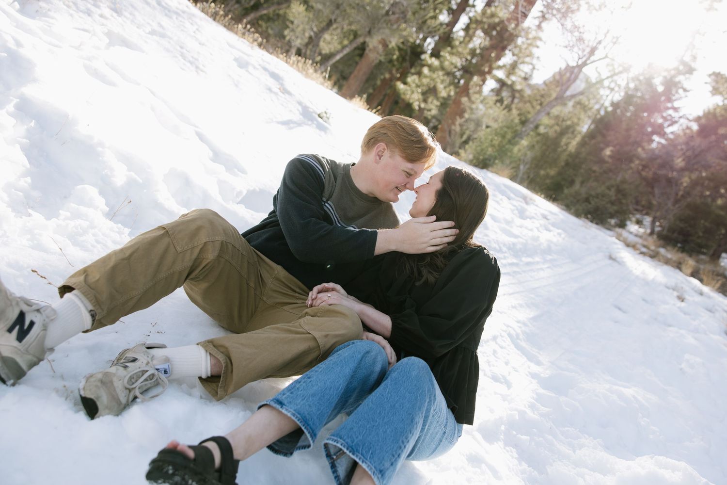 A couple shares a romantic moment lying in the snowy ground against a wintry forest backdrop.