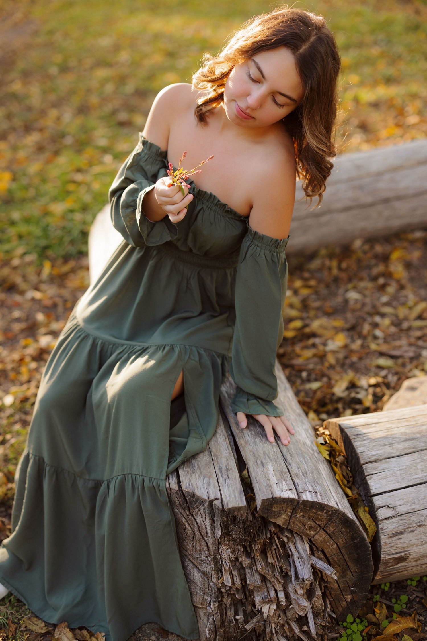 A woman in an olive green off-shoulder dress sits on a fallen log surrounded by autumn foliage at golden hour.