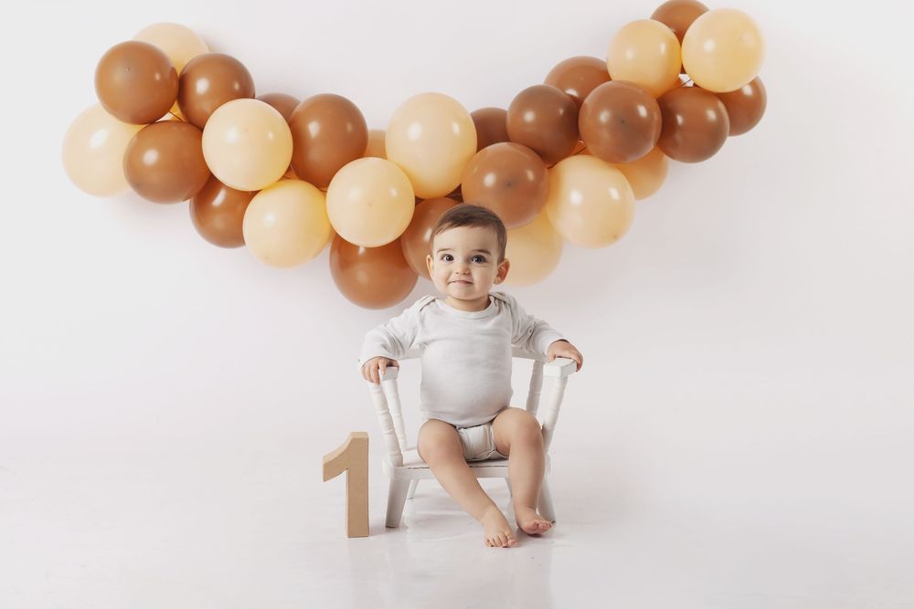 Baby in grey onesie sits on white backdrop surrounded by brown and cream colored balloons for first birthday photos.