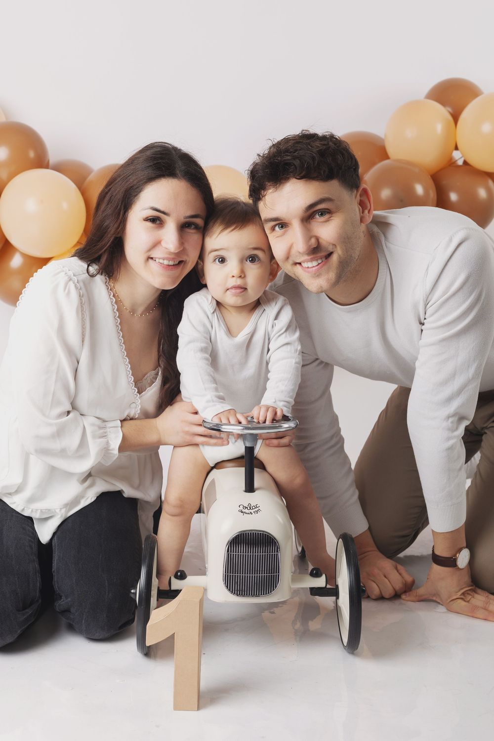 A family in matching white tops poses together against a wall with peach and brown balloons for a photo series.