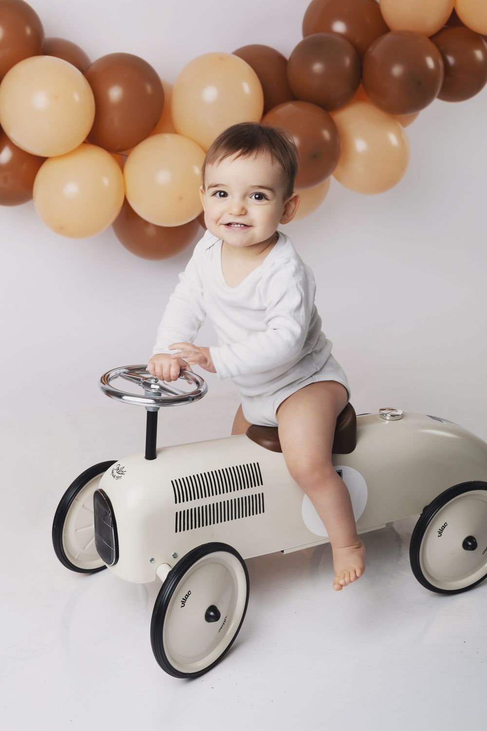 A vintage-style white ride-on toy car against a backdrop of brown and beige balloon decorations.