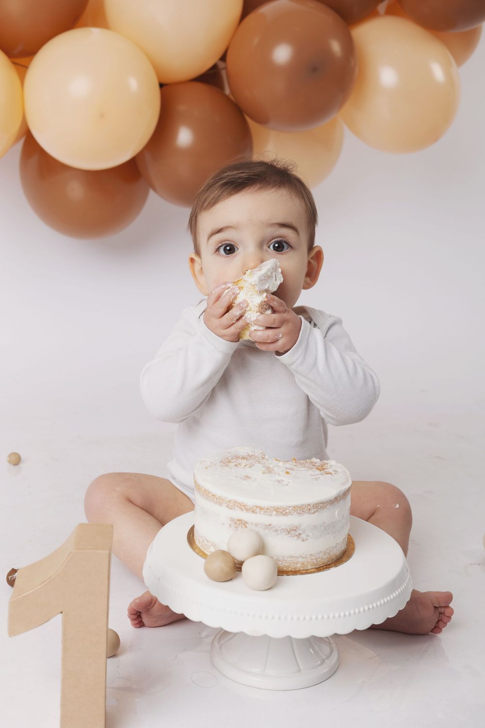 Baby in white outfit sits near a smash cake with brown and beige balloon garland backdrop for first birthday celebration.