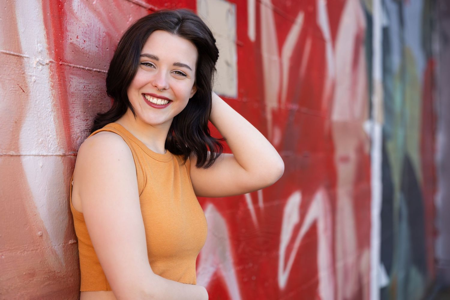 Someone wearing a yellow sleeveless top smiles while leaning against a wall with red graffiti.