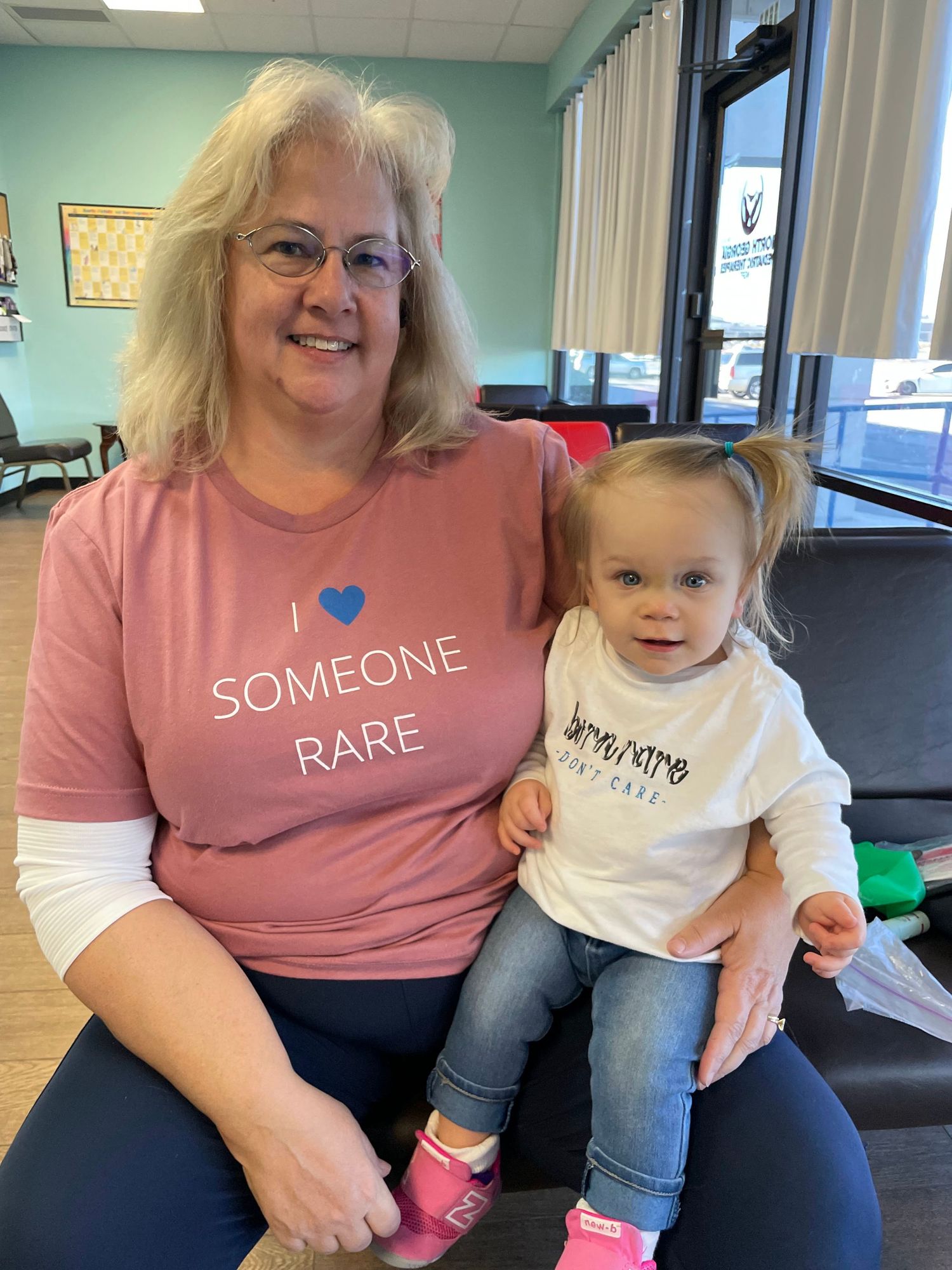 Two people wearing matching 'Someone Rare' themed shirts sit together on a blue bench in a medical office setting.