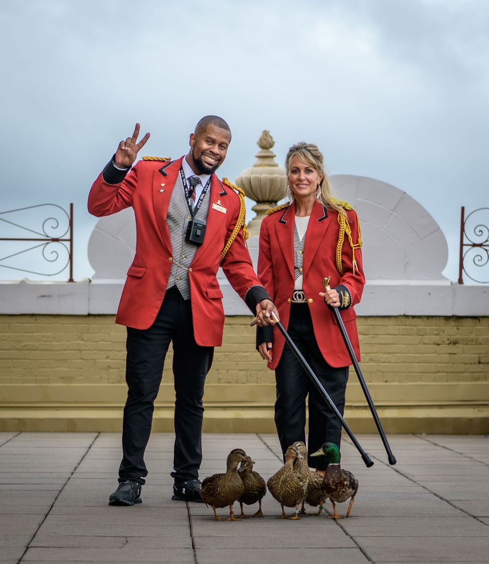 Two tour guides in red blazers standing with ducks on a patio at a historic property during an overcast day.