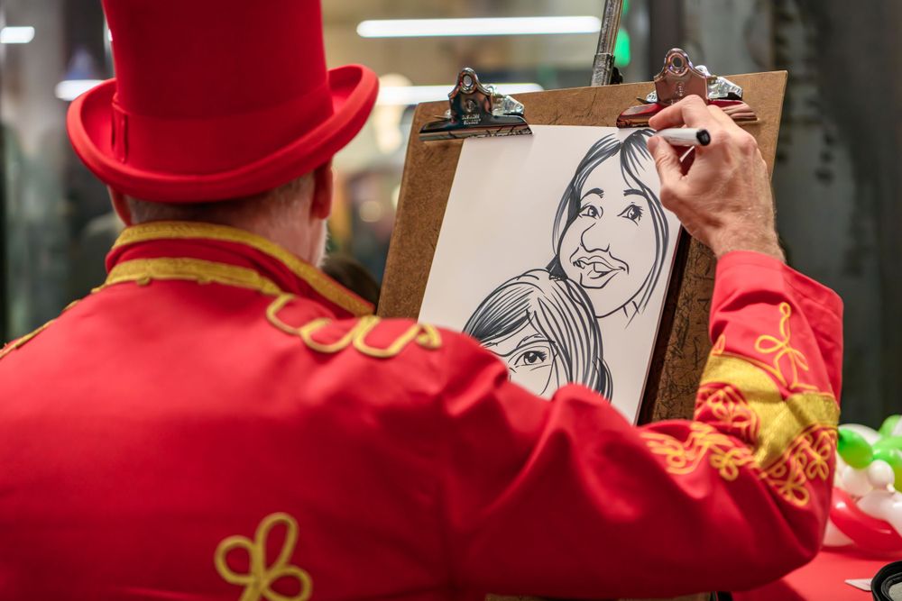 Street artist in red uniform with matching top hat drawing a sketch on a clipboard at an outdoor event.