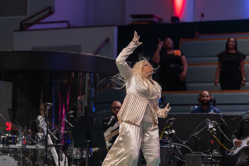 A performer in cream-colored attire gestures dramatically on stage during a live musical performance.