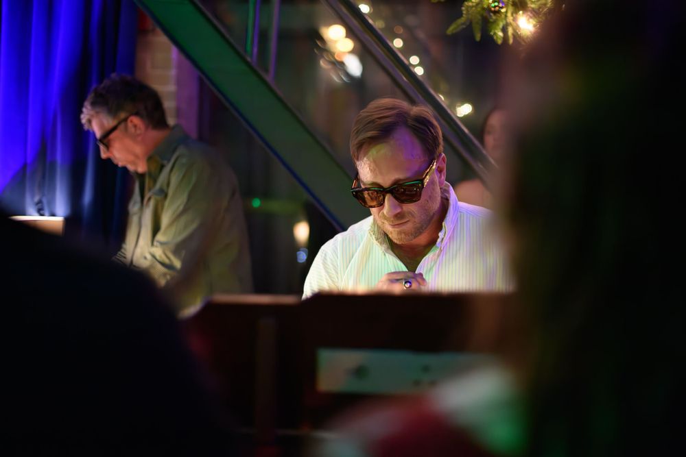 Musicians perform at night under colored stage lighting with one performer wearing sunglasses in a dim venue.