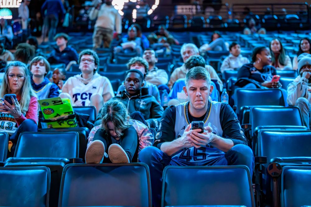 Audience members sit in blue stadium seats looking at mobile phones and devices during an event.
