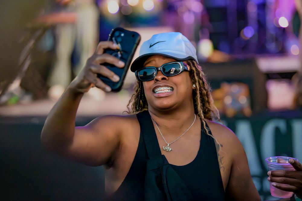 A concert-goer in sunglasses and a white cap takes a selfie while holding a drink in a dark venue with purple lighting.