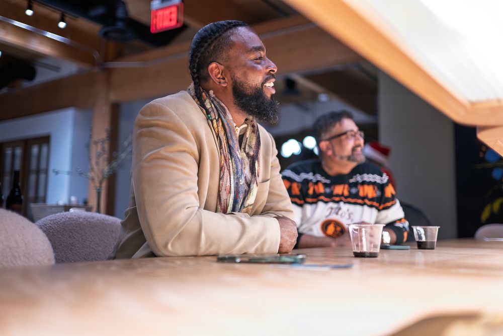 A person in a beige suit jacket sits at a bar counter while laughing and enjoying a beverage.