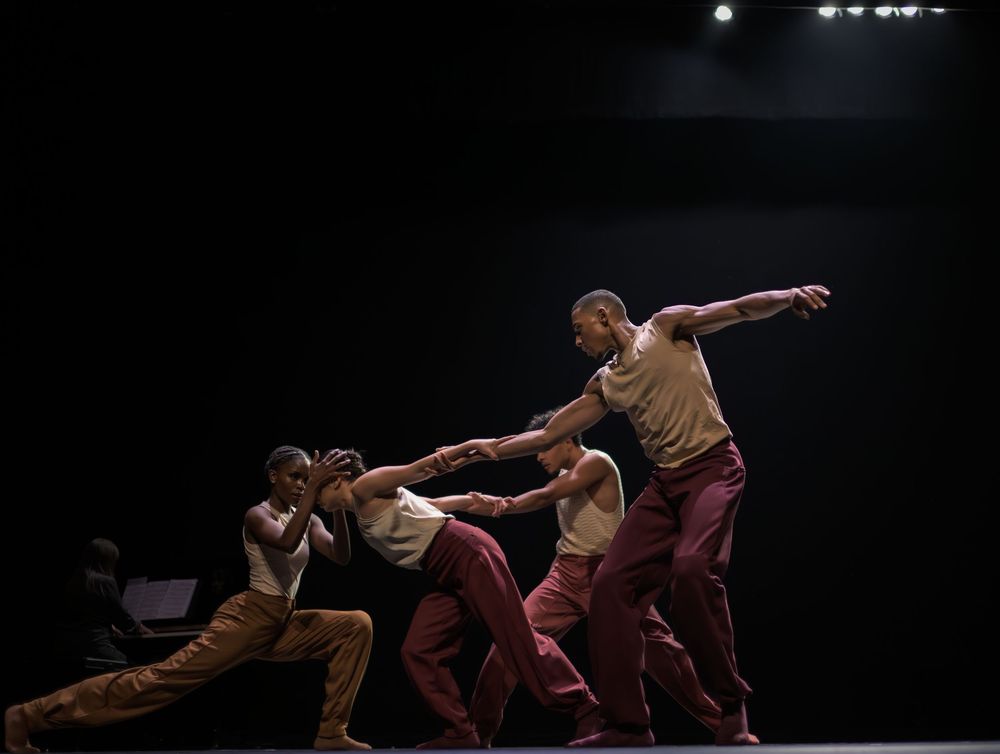 Modern dancers in brown and burgundy costumes perform a dynamic sequence of movements on a dark stage.
