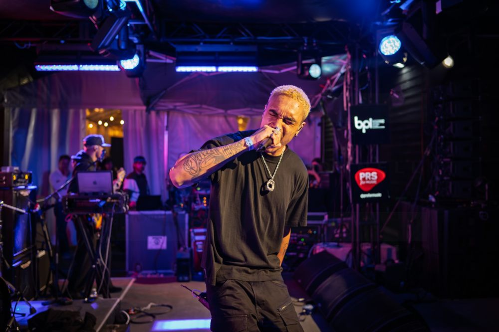 Tattooed male performer with blonde hair sings on stage under colorful lights at an indoor music venue.