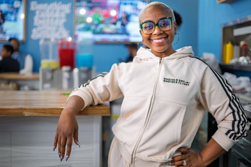 A person wearing glasses and a cream-colored hoodie smiles while leaning against a counter in a casual dining setting.