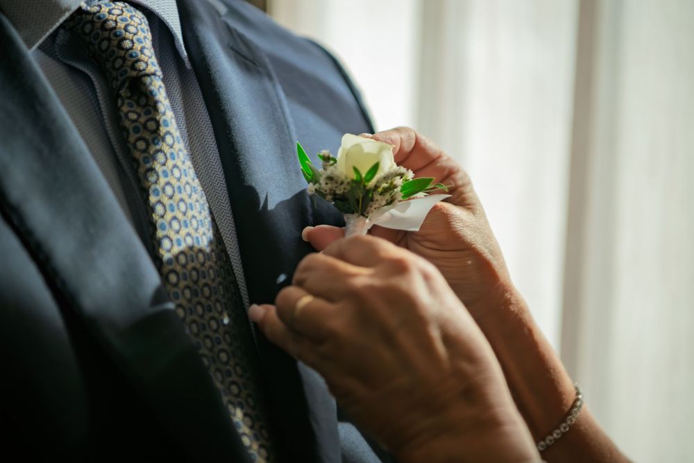 Hands pin a white rose boutonniere with green leaves onto the lapel of a man in a navy suit and patterned tie at a wedding.