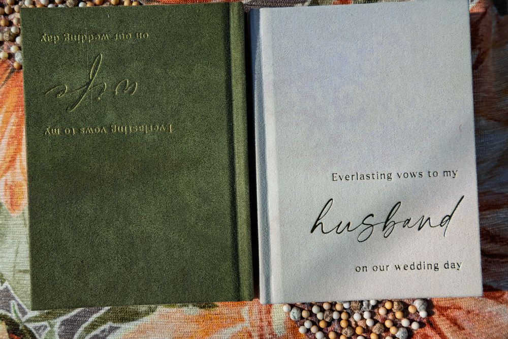 Two linen-bound wedding vow books, green for wife and light blue for husband, with gold lettering, resting on a floral fabric.