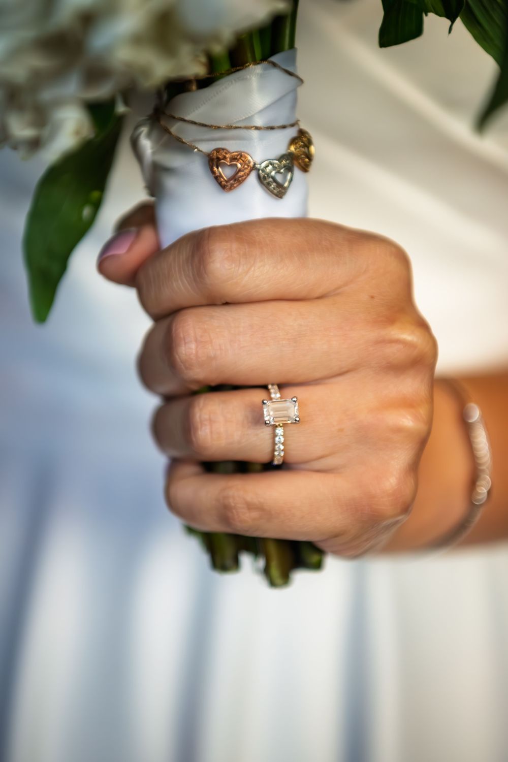 Bride's hand wearing an emerald-cut diamond engagement ring holds a bouquet wrapped in white ribbon with heart-shaped charms.