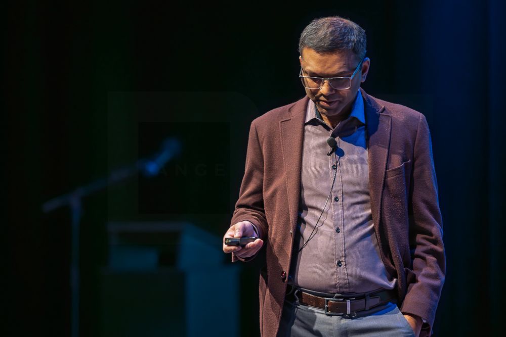 Speaker wearing a brown blazer and light colored shirt gestures while presenting on stage at a conference event.