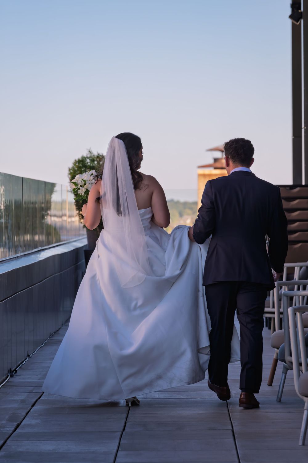 Bride in a white gown and veil walks with her groom in a dark suit along a rooftop terrace at sunset, carrying a flower bouquet.
