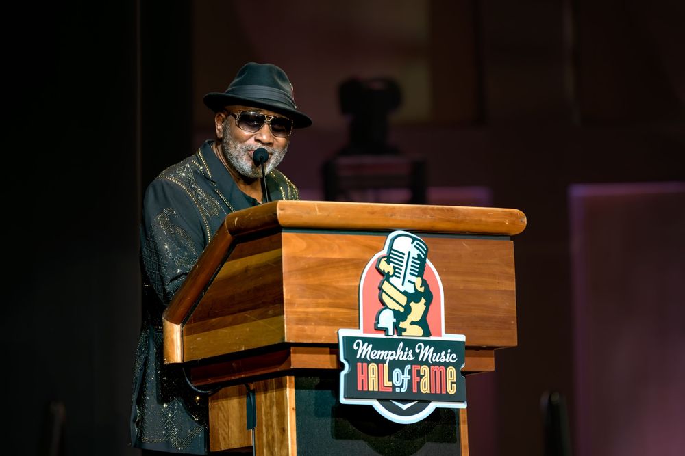 A person wearing a hat and sunglasses speaks at a wooden podium with a sports team logo visible.