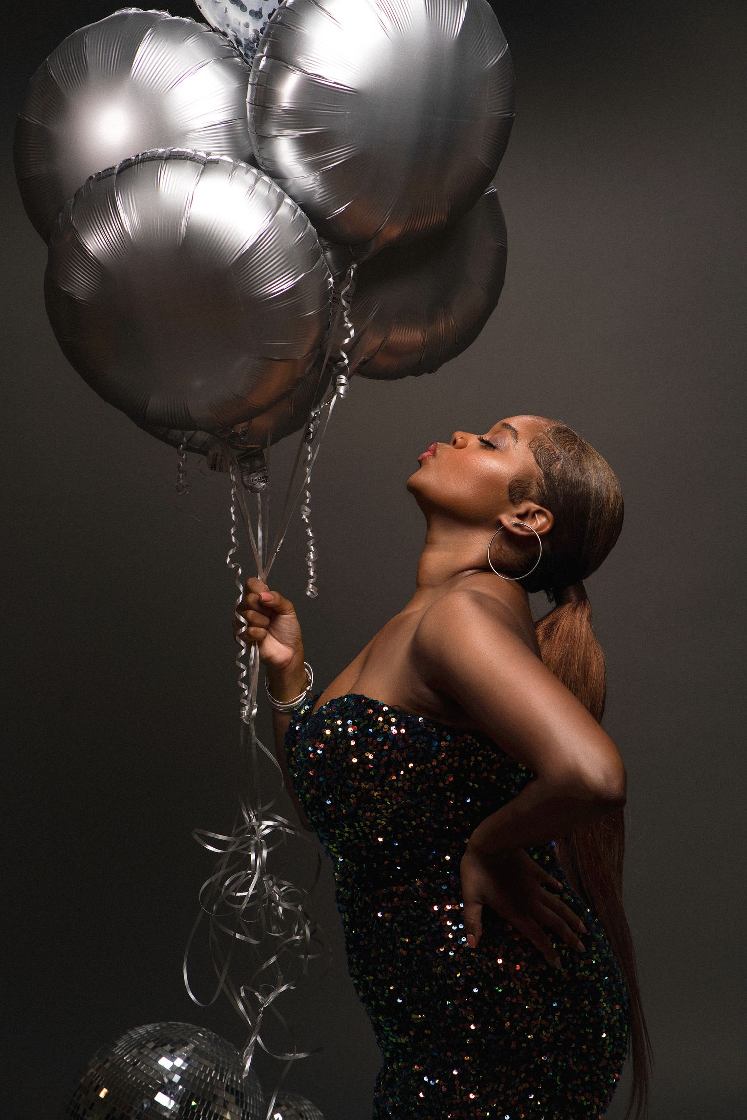 Woman in a sparkly black dress poses elegantly with silver metallic balloons against dark background.