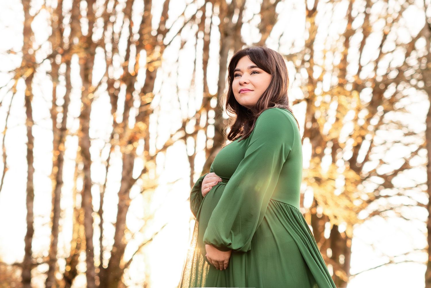 Someone in a emerald green dress smiles while standing in a wooded area with autumn trees in the background.