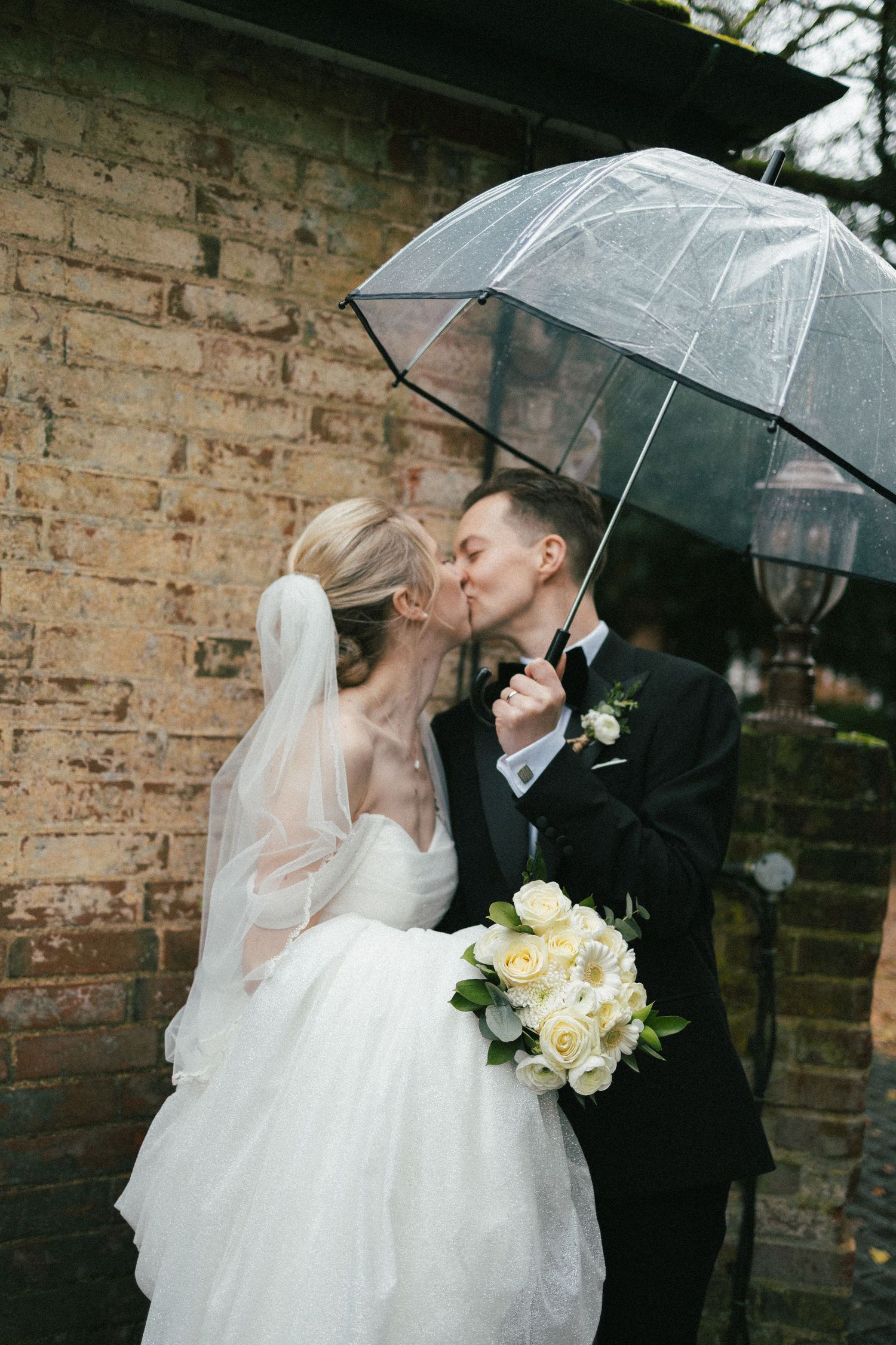 Romantic wedding couple sharing a moment under a clear umbrella against brick wall.