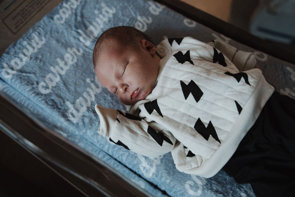Newborn baby sleeping peacefully on blue blanket wearing white sleeper with black bow pattern.