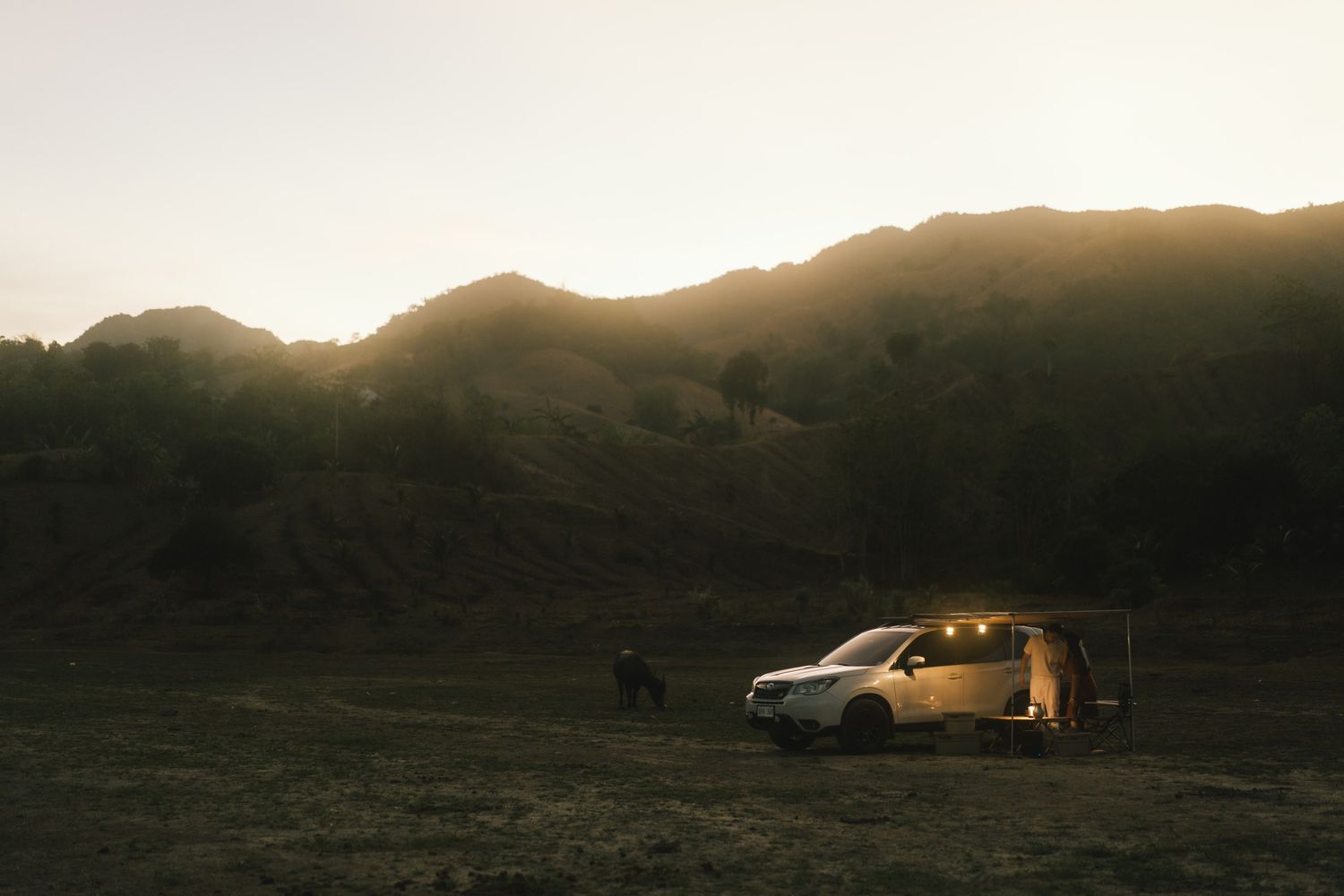Car parked on hillside against dramatic mountain silhouette at sunset.