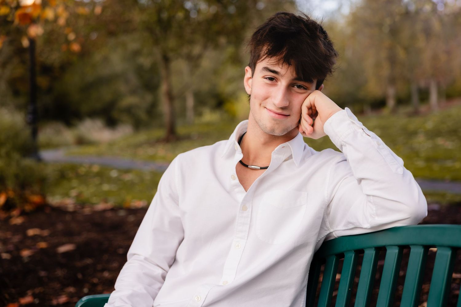 A person in a white button-down shirt sits casually on a green park bench during autumn.