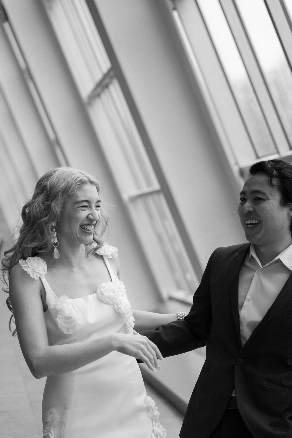 A joyful couple shares a candid laughing moment in a bright setting with angled windows during their wedding day.