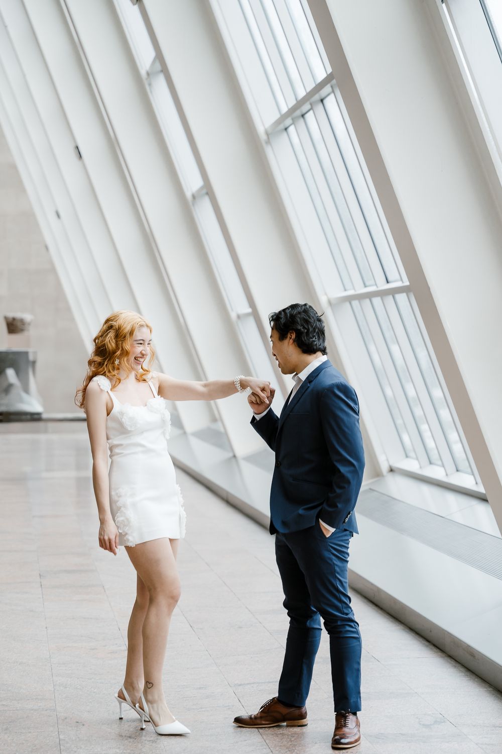 An intimate gesture between two people standing in a bright hallway with dramatic architectural windows.
