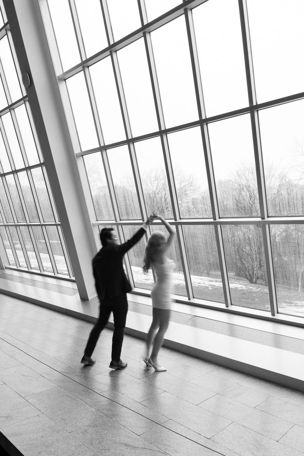 A couple dances together in front of large angled windows in a modern building, captured in black and white.