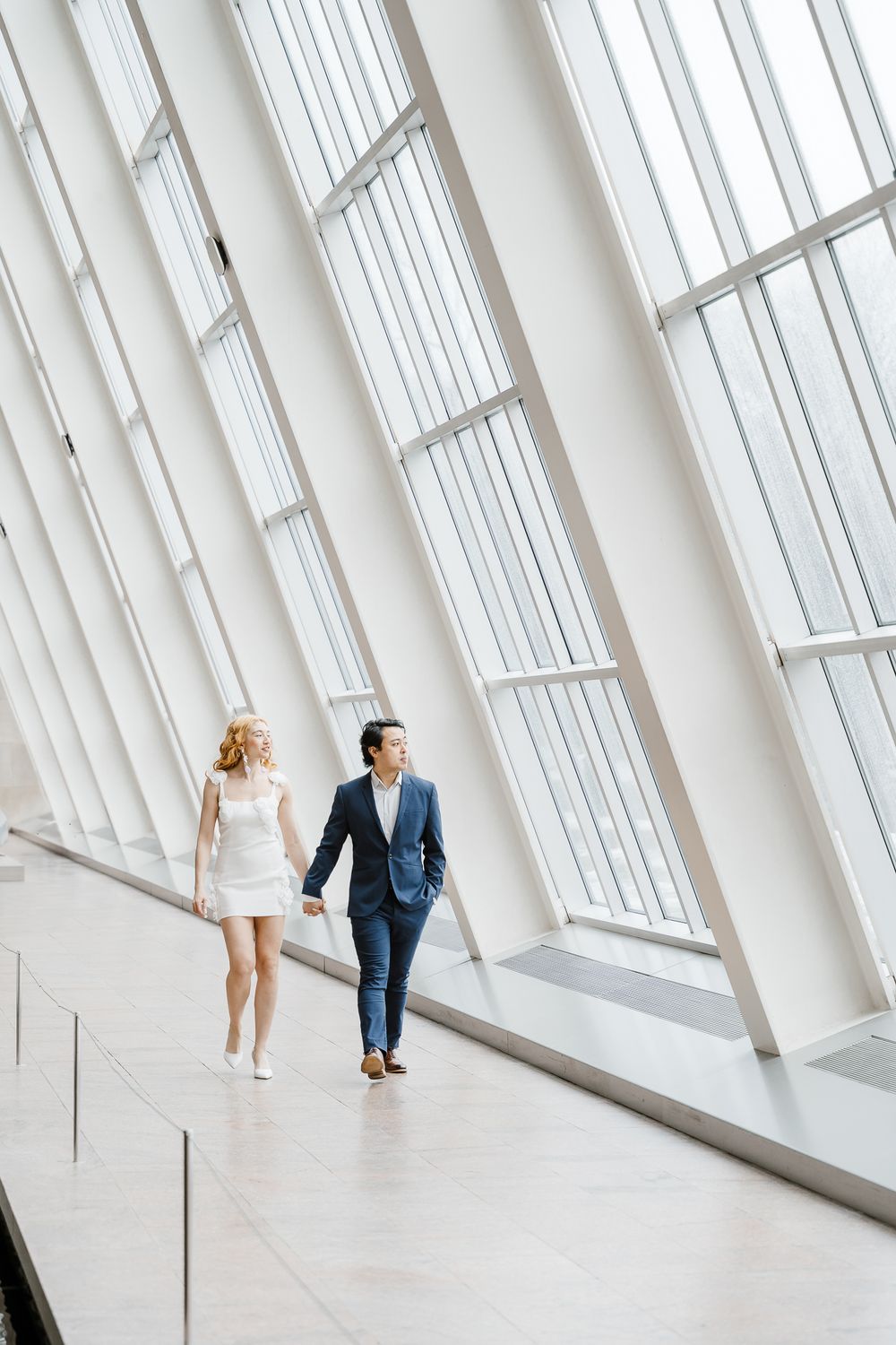 A couple walks hand in hand through an elegant modern building with dramatic angled windows and white architecture.