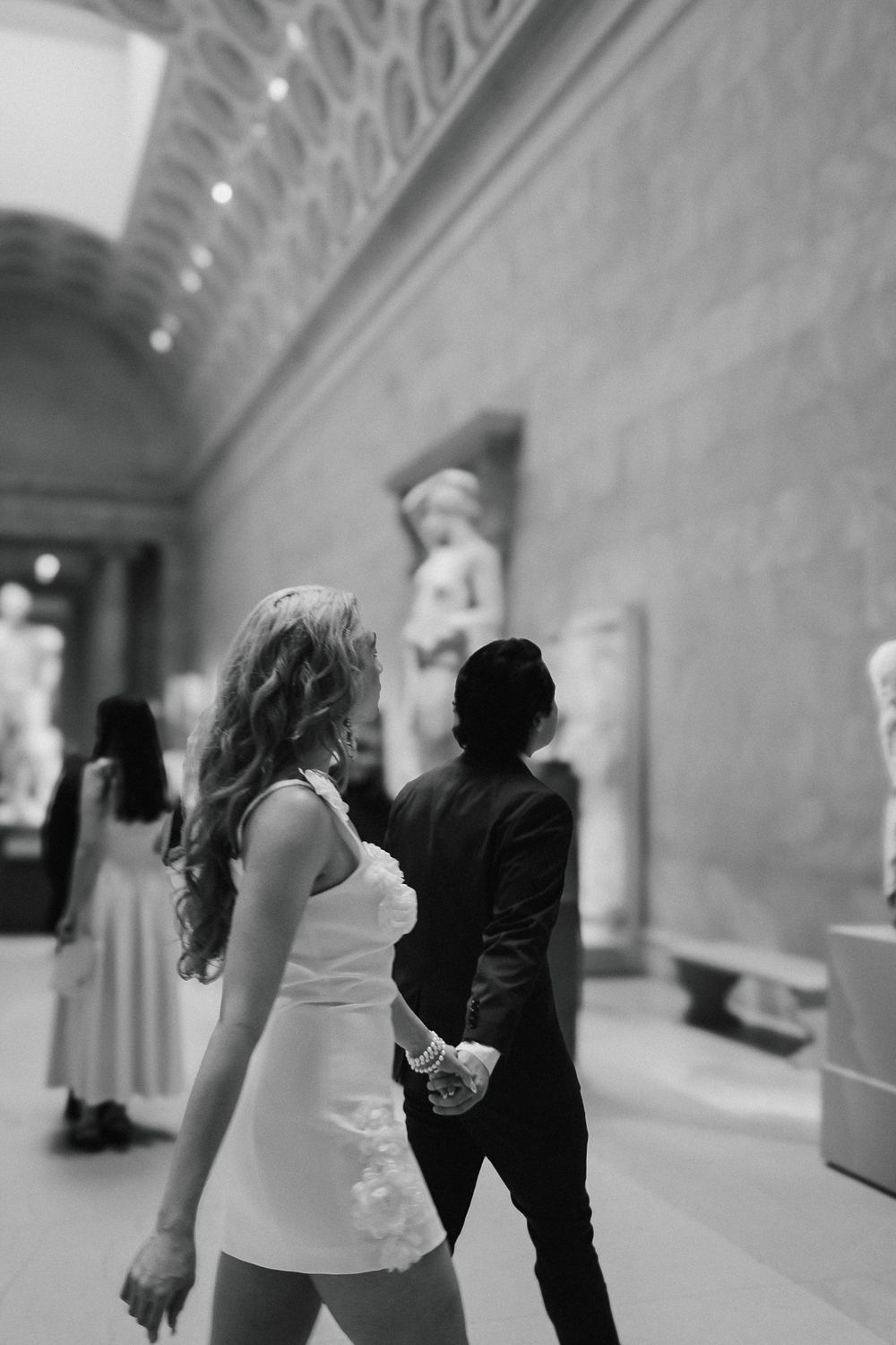 A couple walks through an art museum gallery featuring classical statues and ornate architecture.
