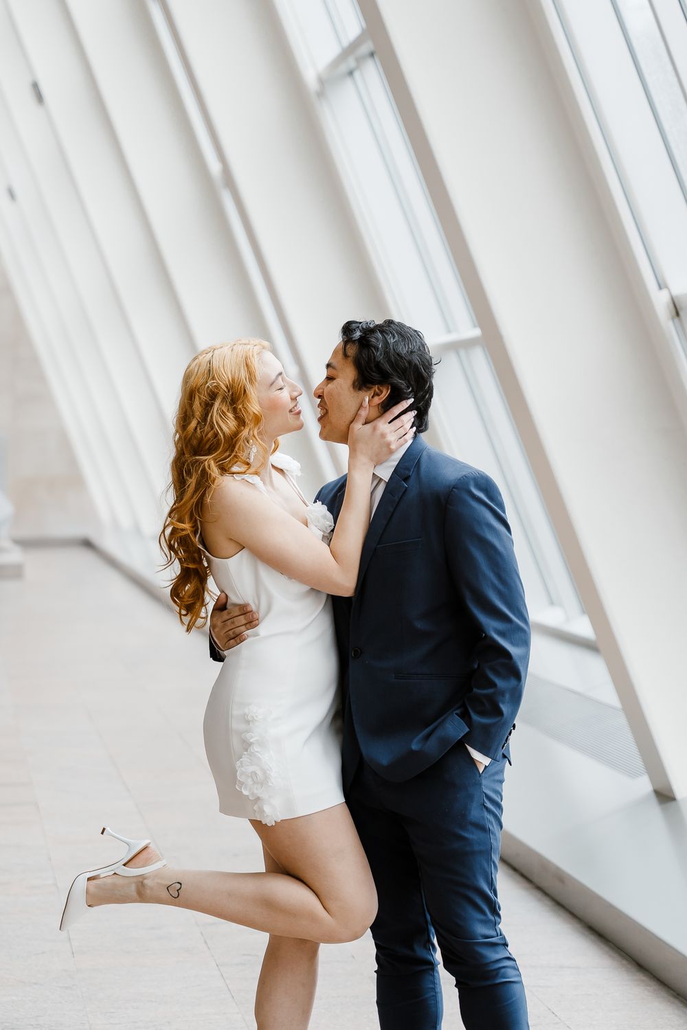 A romantic couple embraces in a bright modern hallway with angled white walls and natural lighting.