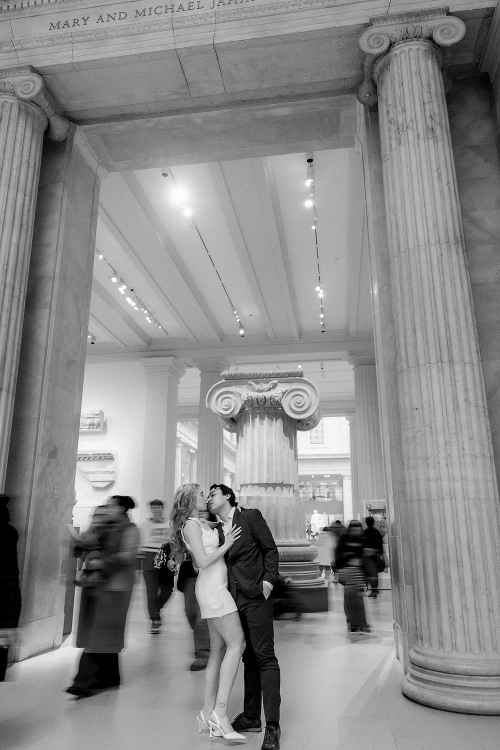 A romantic moment between a couple in a museum atrium with classical columns and passing visitors.