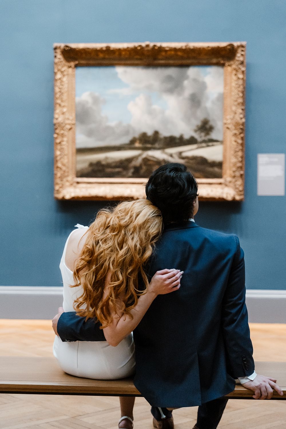 A couple sits close together on a bench while admiring a framed landscape painting in an art museum.