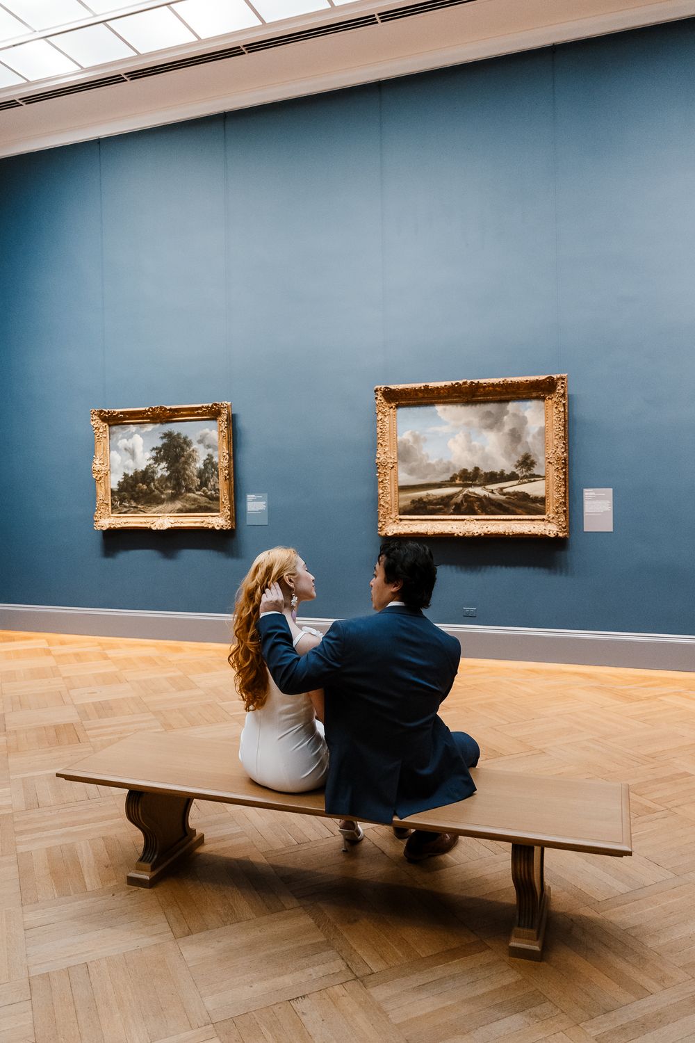 Two visitors sit on a wooden bench viewing landscape paintings on a blue wall at an art museum.