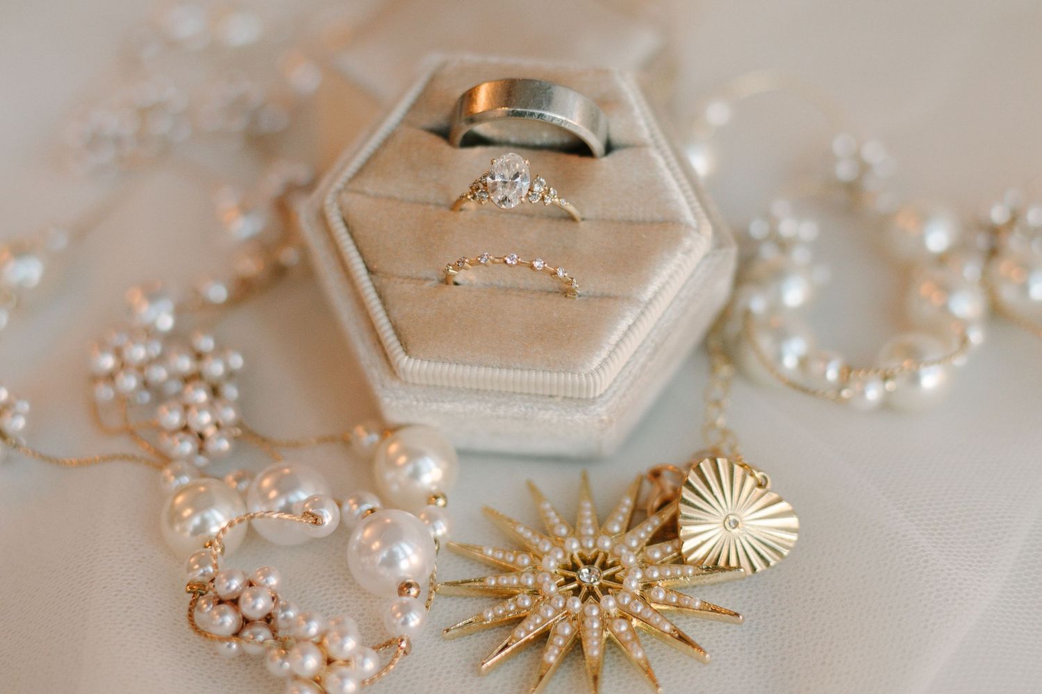 Wedding jewelry including a diamond ring in hexagonal box, pearl necklace, and gold starburst brooch on beige background.
