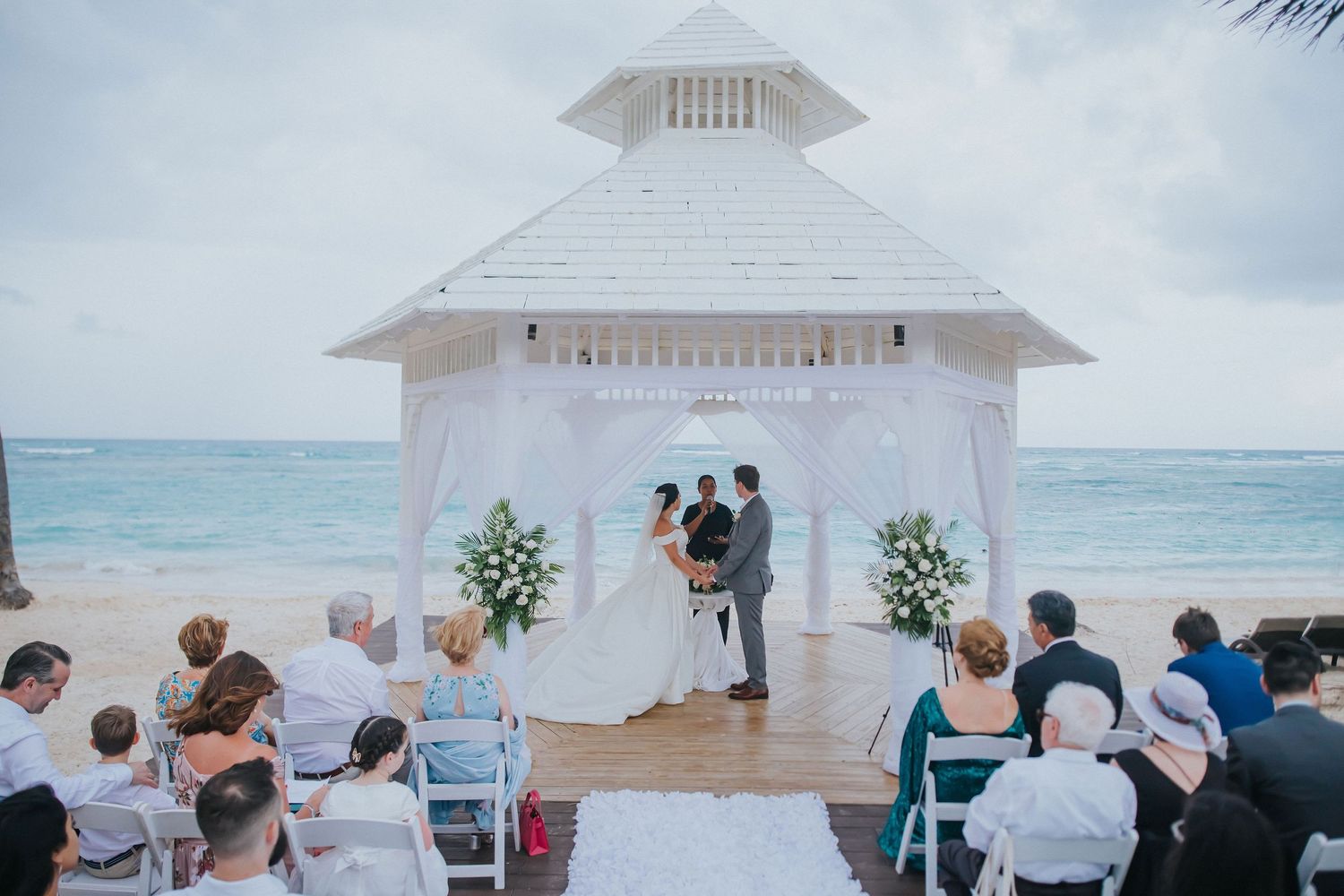 Beachfront wedding ceremony at a white gazebo with ocean views and seated guests.