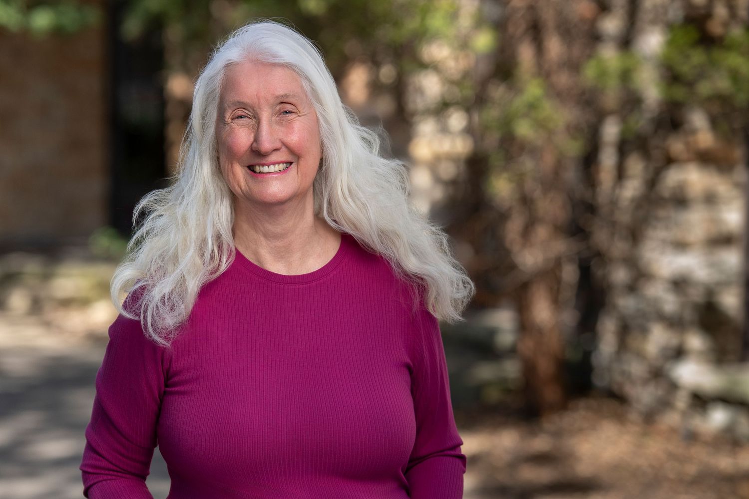 A person with long silver hair wearing a magenta sweater smiles outdoors on a sunny day.
