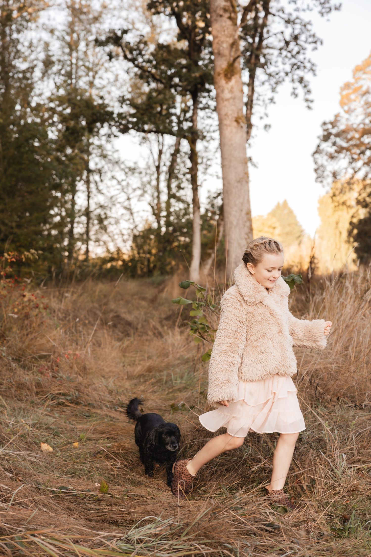 Child in fluffy cream coat walking through autumn woods with small black dog in golden sunlight.