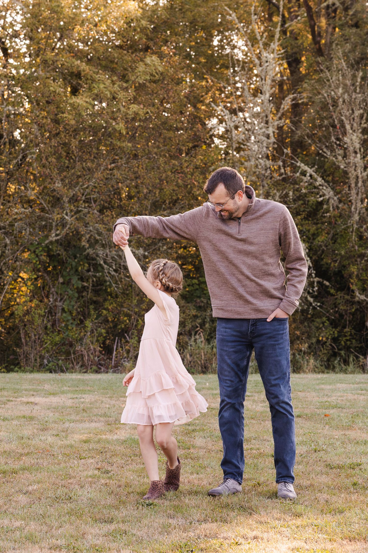 A child in a pink dress dances with an adult wearing a brown sweater in an autumn field at sunset.