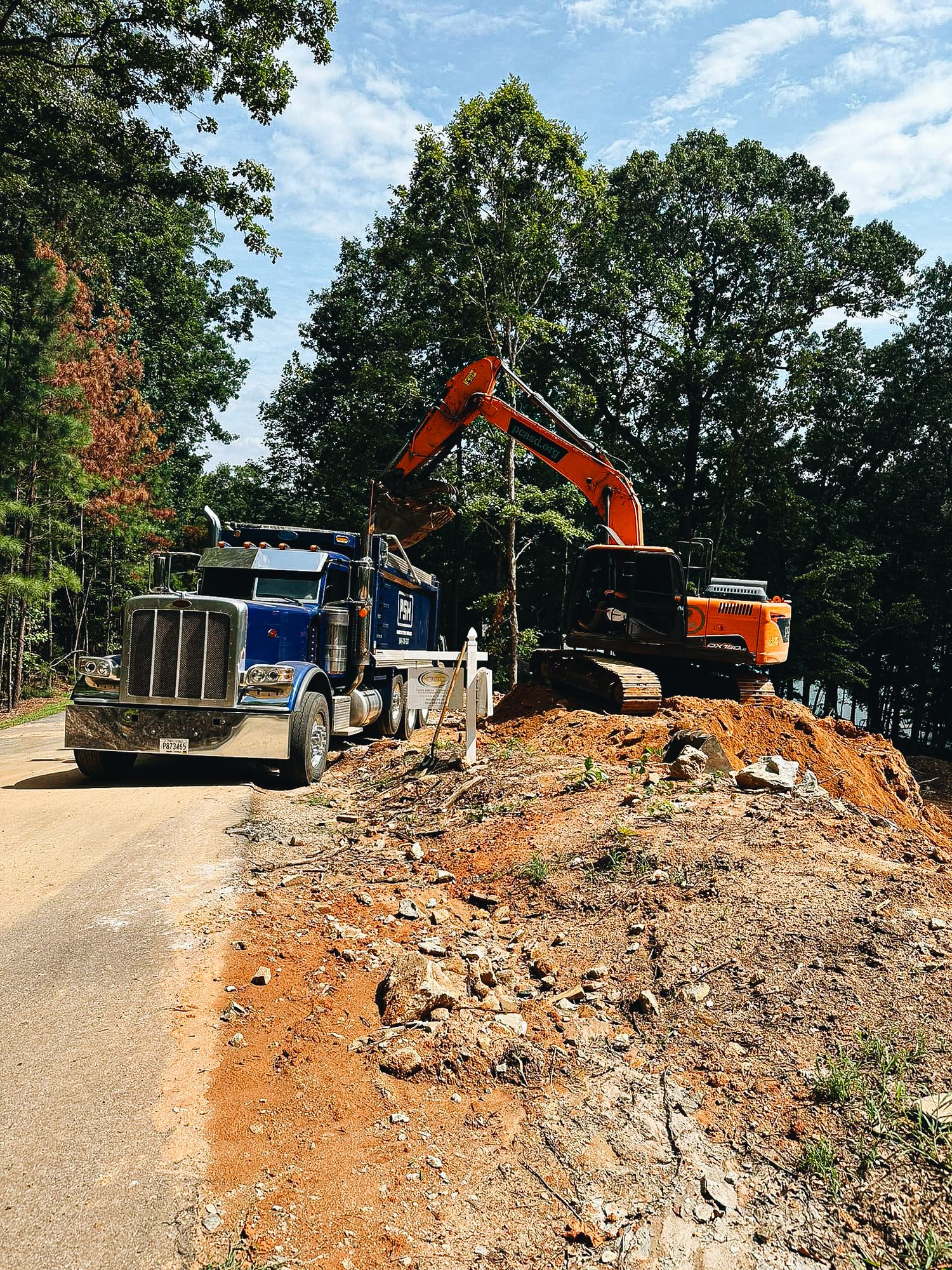 Construction excavator loads dirt and debris into dump truck during roadwork on rural forested road.