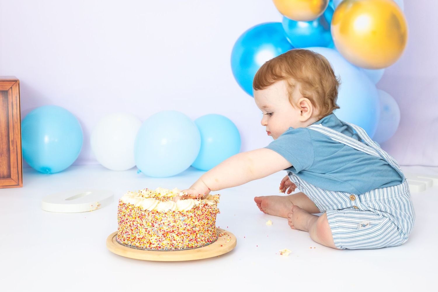 Baby in blue outfit reaching towards a decorated birthday cake surrounded by festive balloons.