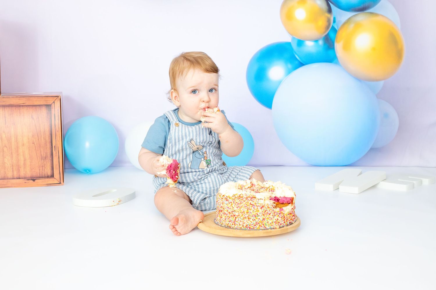 First birthday celebration scene with blue and gold balloons and a decorated cake on white background.