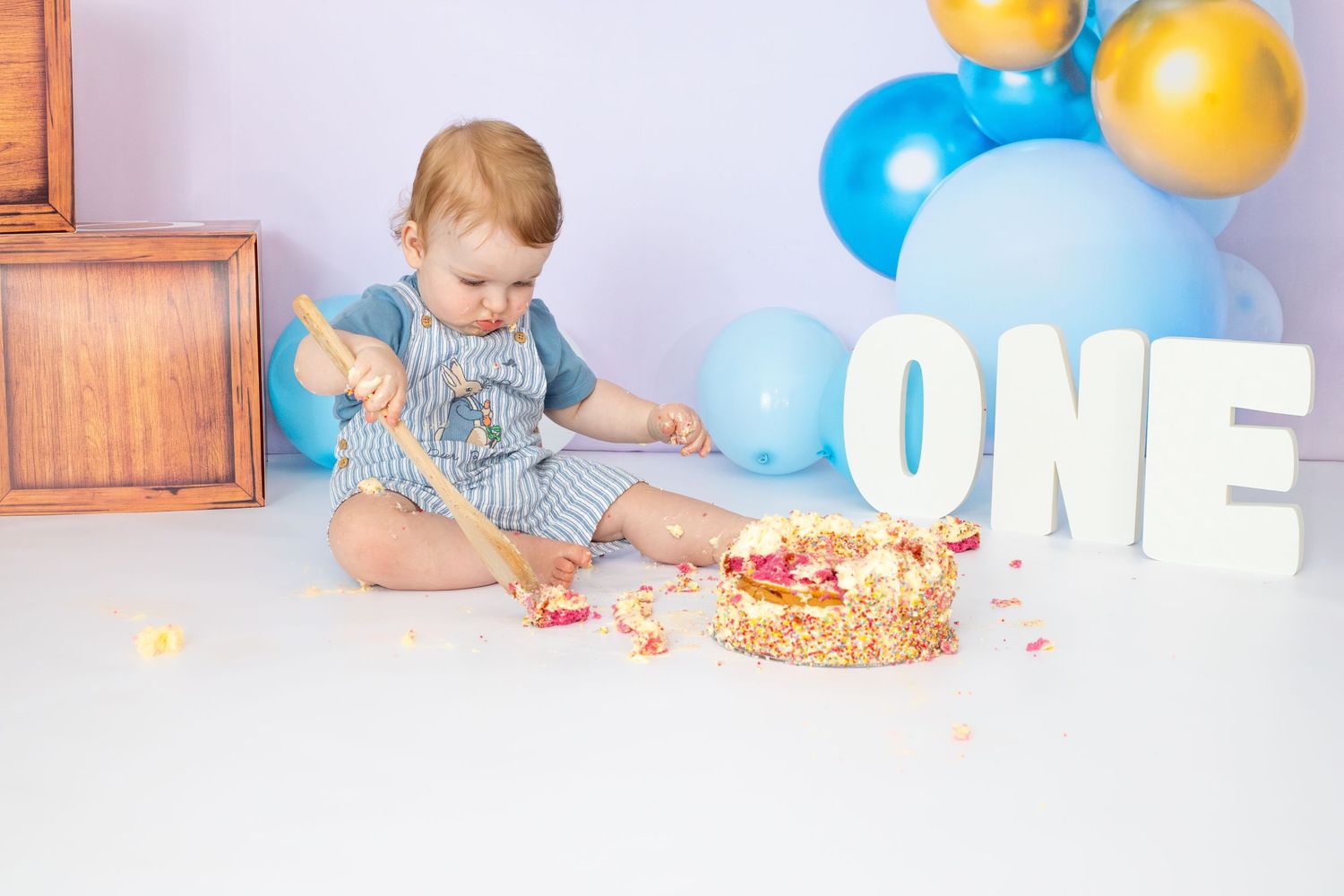 Birthday setup with 'ONE' letters, balloons, and cake against white backdrop.